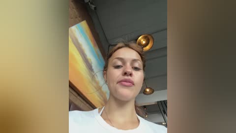 Video of a young Caucasian woman with light skin, brown hair, and wearing a white t-shirt, standing in a modern, well-lit room with a colorful, abstract painting on the wall and round pendant lights.