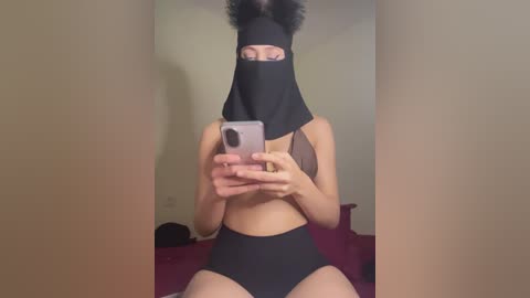 Video of a slim, light-skinned person with curly hair, wearing a black face mask, black top, and high-waisted black shorts, holding a phone. Background shows a plain wall and red bedding.