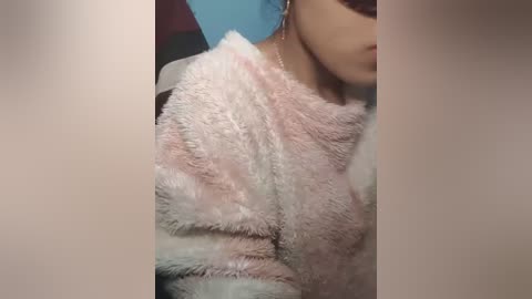 Video of a young woman with light skin, wearing a fluffy, pastel-colored jacket, and a delicate necklace. She is partially visible, with her face cropped out. Background is a blurred blue wall.