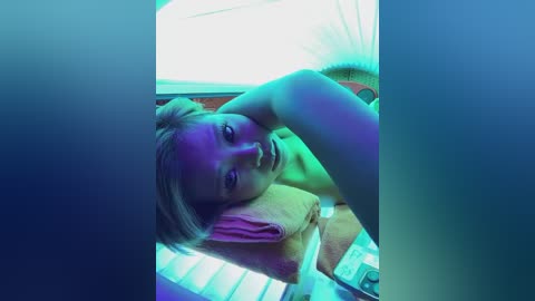 Video of a blonde woman with a towel on her head, lying on a tanning bed with neon blue and purple lighting, giving a relaxed, slightly contemplative expression.