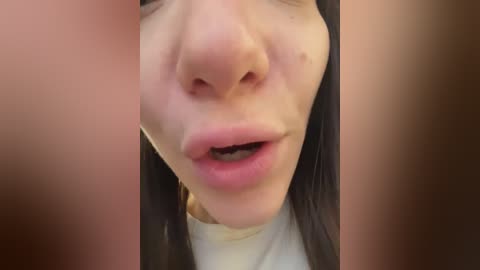 A close-up video of a woman's face, showing her nose, lips, and a portion of her forehead. She has fair skin, long black hair, and is wearing a white top. The background is blurred and out of focus.