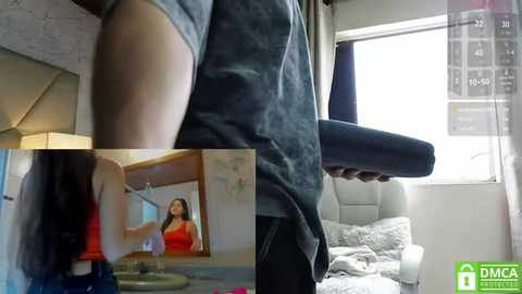 Video of a man in a grey shirt lifting a large black object, while a woman in a red dress stands in a modern bedroom with white walls and a window.