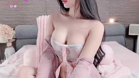 Video of an Asian woman with long black hair, wearing a strapless lace bra and a pink robe, sitting on a bed with a gray headboard and pink sheets, in a modern bedroom.