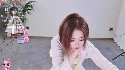 Media: Video of a young East Asian woman with straight, shoulder-length brown hair, wearing a cream sweater, lying on a gray carpet in a minimalist room with a white wall and a green plant in the background.