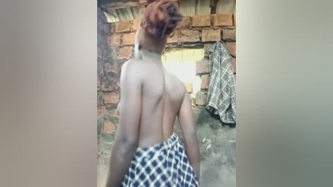 Video of a shirtless man with fair skin and short brown hair, wearing blue and white plaid shorts, standing in a rustic, brick-walled room with a hanging shirt.