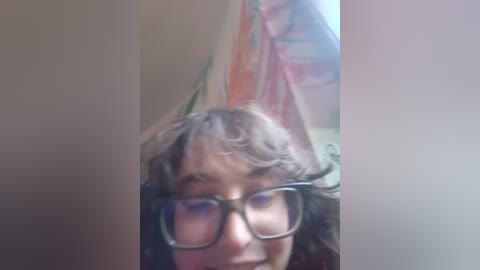 A close-up video of a person with short, wavy gray hair and glasses, wearing a dark shirt. The background shows a blurred, colorful wall with abstract patterns. The image has a slightly blurry quality.