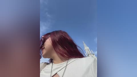 Video of a young woman with straight, long red hair, wearing sunglasses, a white ribbed top, and a silver necklace, looking upward against a clear blue sky.