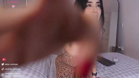 Video of a Latina woman with long dark hair, wearing leopard print lingerie, sitting on a bed, blurred background, social media icons in the corner.
