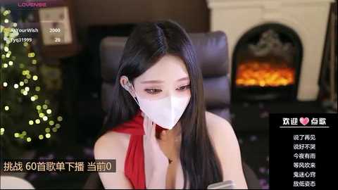 Media: A video of an East Asian woman with long black hair, wearing a white face mask, red dress, and earphones, sitting indoors near a lit Christmas tree and fireplace.
