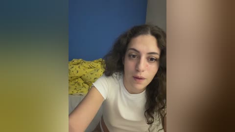 Video of a young woman with long, curly dark hair and fair skin, wearing a white t-shirt, leaning against a blue and yellow wall.