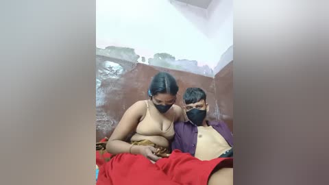 Video of a young woman in a beige top and black mask, sitting on a bench in a dimly lit, rustic bathroom with a red towel draped over her lap. A young man in a purple shirt sits beside her.