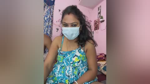 Video of a South Asian woman with medium-dark skin, wearing a blue surgical mask, colorful floral dress, and black hair tied back. She sits in a pink-painted room with a blue curtain and colorful decorations.