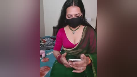 Video of a South Asian woman with long black hair, wearing a black face mask, red top, and green sari, sitting on a bed with a colorful blanket, holding a smartphone.