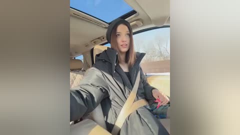 Video of a young woman with straight brown hair, wearing a black beanie and puffy gray coat, seated in a car with beige seats, sunlight streaming in, and a clear blue sky visible through the window.
