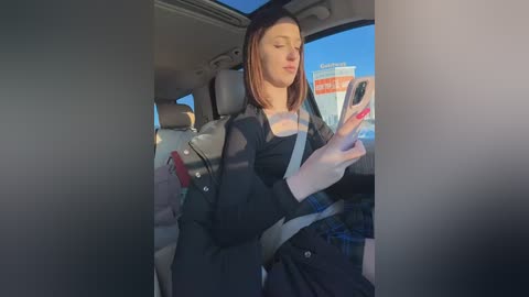Video of a woman with light skin, brown hair, and a black jacket, taking a selfie in a car. The background shows a clear blue sky and a white building.