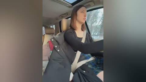 Video of a young woman with shoulder-length brown hair, wearing a black coat, driving a car with a plaid skirt visible in the background.