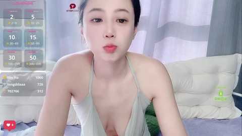 Video of an Asian woman with pale skin, dark hair, and a small bust, wearing a low-cut white top, blowing a kiss in a bedroom with white curtains and a bed.