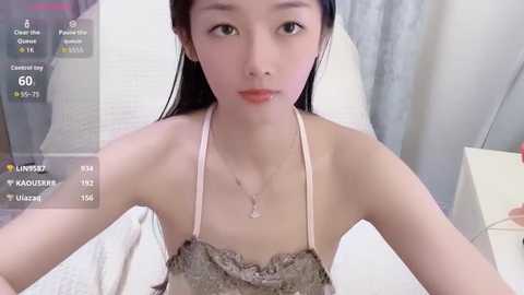 Video of a young East Asian woman with long black hair, wearing a light pink lace camisole, lying on a white bed, surrounded by a modern bedroom setting.