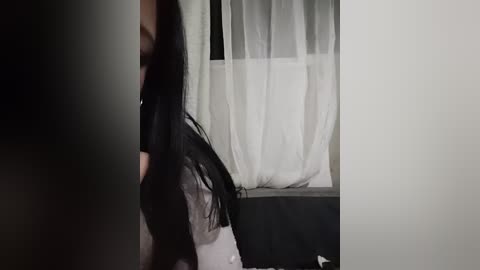 Video of a person with long black hair, wearing a black top and grey pants, standing near a window with white curtains, dim lighting.