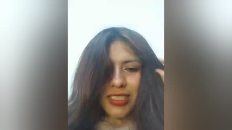 Video of a young woman with long, dark hair, light skin, and red lipstick, smiling. She has one hand holding her hair aside, revealing her face. Background is blurred, focusing on her expression.