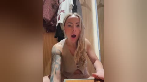 Video of a young woman with long blonde hair and tattoos, wearing a beige tank top, sitting in a locker room. She looks surprised, mouth open, with a towel and clothes hanging nearby.