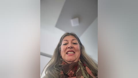 A video of a smiling, middle-aged woman with long, blonde hair, wearing a floral-patterned blouse, standing in a modern, white room with a triangular ceiling light fixture.