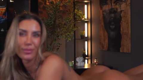 Video of a blonde, topless woman with fair skin and medium breasts, lying face down on a bed in a dimly lit room with modern decor, including a leafy plant, a black bust sculpture, and abstract art.