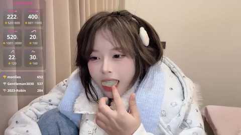 Video of an East Asian child with straight, shoulder-length brown hair, wearing a white puffy jacket with a cartoon whale pattern. They are licking a lollipop, with a digital overlay showing live stream stats.