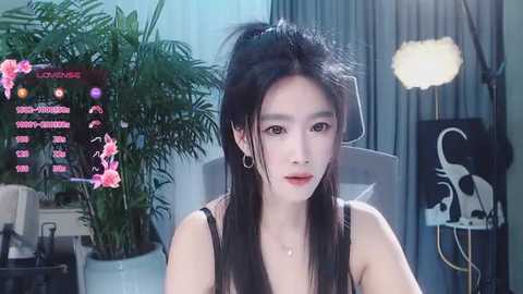 Media: A video of a young East Asian woman with long black hair in a ponytail, wearing a black top, sitting in a room with a green plant, a screen displaying chatroom messages, and a studio light.