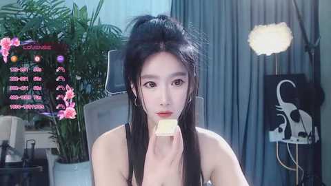 Media: Video of an East Asian woman with pale skin, black hair, and a slender physique, eating a slice of cake. Background includes a green plant, a gray chair, and a lighted studio setup.