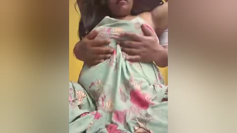 A video of a young girl with medium brown skin, wearing a floral-patterned dress, holding her chest, partially covered by her hands, against a yellow wall.