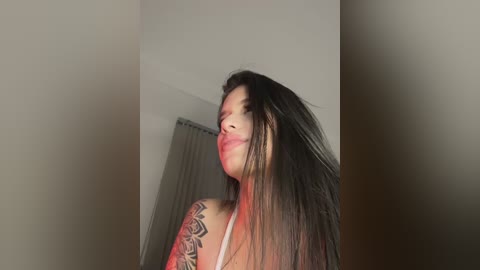 Video of a woman with long, straight black hair, wearing a white top, against a plain, beige wall with a partially visible window. Her face is slightly tilted to the side, and her lips are pursed.