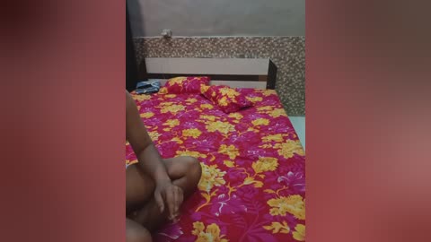 Video of a naked, dark-skinned person sitting on a bed with a red and yellow floral quilt, in a small, dimly-lit room with brown and beige walls.