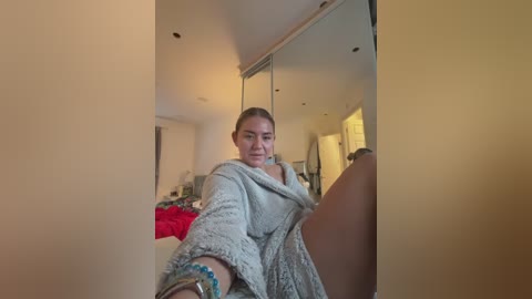 Video of a young woman with fair skin and blonde hair, sitting on a bed in a modern, brightly lit bedroom, wearing a grey, textured robe. The background shows a white wall and a large mirror.