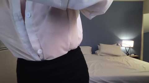 Video of a person adjusting a light blue dress shirt, standing in a dimly lit bedroom with white bed, blue wall, and bedside lamp.