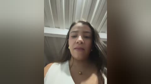 Video of a young woman with long black hair, light skin, wearing a white sleeveless top, standing indoors with a white ceiling and light wooden walls.