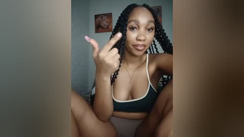 Video of a young Black woman with braided hair, wearing a black sports bra, sitting on a bed, making a middle finger gesture.