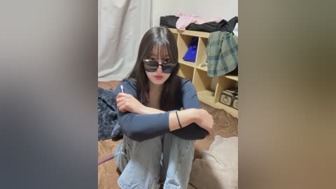Video of an Asian woman with long black hair, wearing sunglasses, a blue long-sleeve shirt, and jeans, sitting on a carpeted floor in a cluttered room.