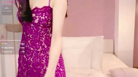 Media: Video of a woman in a revealing, purple lace lingerie dress, standing in a modern bedroom with light pink walls and white bedding.