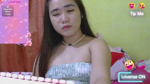 A video of a young Asian woman with long black hair, wearing a strapless, patterned top, lying on a bed with a pink pillow, in a room with yellow walls and a pink heart-patterned wall.