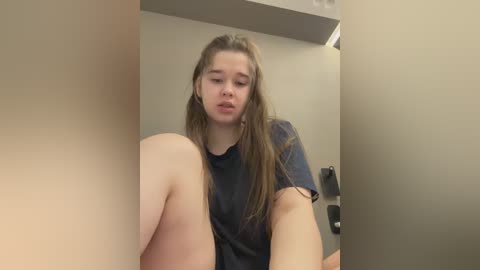 A video of a young Caucasian woman with long brown hair, wearing a dark t-shirt, sitting on a beige floor. The background is a plain, beige wall with a doorframe and a ceiling light.