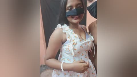 Video of a young woman with dark skin, wearing a white floral dress, black face mask, and black hair, standing in an indoor setting.