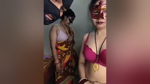 Video of three women in Indian attire: one in a brown sari with a red blouse, another in a red sari with a gold necklace, and a third in a pink bra with a golden pendant.