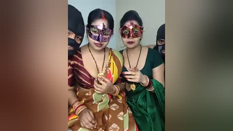 Video of two women in traditional Indian attire, wearing colorful, ornate masquerade masks and beaded jewelry, standing close together against a plain background.