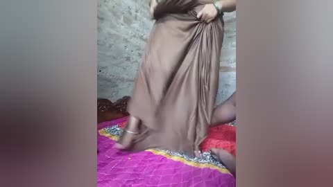 Video of a woman in a long, flowing brown dress, lifting the hem, revealing a purple and gold patterned rug, against a rustic brick wall backdrop.