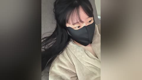 Video of an Asian woman with long black hair, wearing a black face mask, light beige bathrobe, and sitting on a grey couch. Her expression is neutral, and the background is blurred.