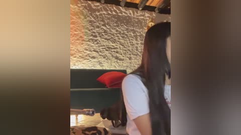 Video of a woman with long black hair, wearing a white t-shirt, partially visible from the side, standing in a rustic room with exposed stone walls, a green couch, and a red pillow.