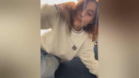 Video of a young woman with wavy, shoulder-length brown hair, wearing a cream sweater with a black emblem and light blue jeans, leaning forward with a playful expression.