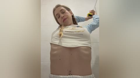 A video of a young, fair-skinned woman with long blonde hair, wearing a white crop top and white pants, holding a yellow bottle, in a small, tiled shower stall.