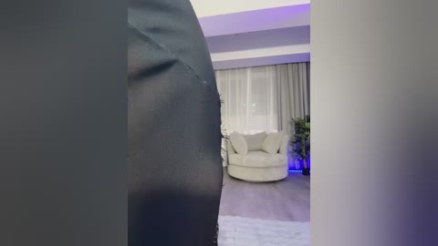 Video of a modern living room with a focus on a person wearing black leggings, standing near a white armchair and a potted plant in the background.